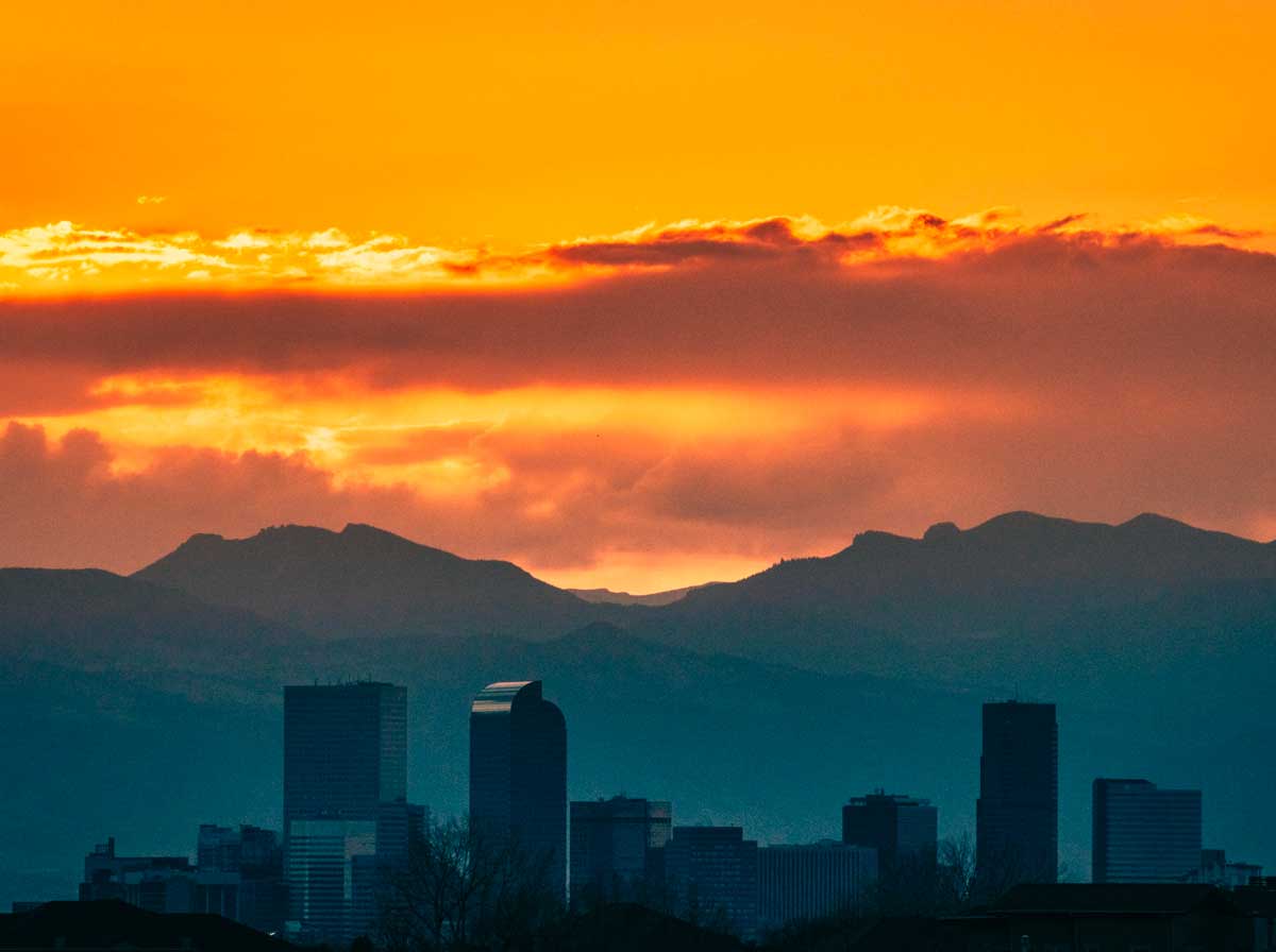 Denver skyline and mountains backlit by a sunset.