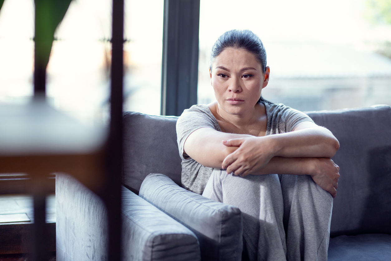 A somber woman sitting on a sofa hugging her knees.