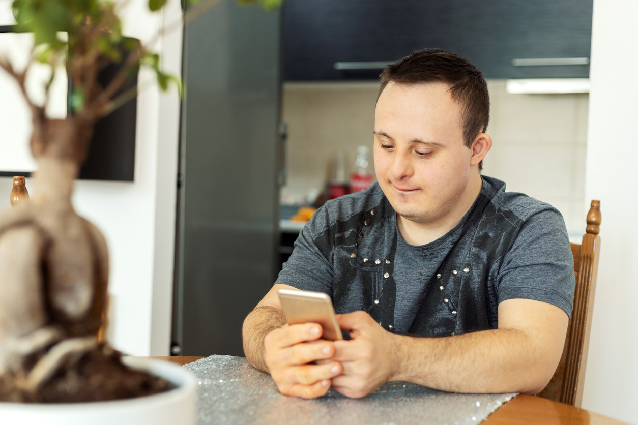 A young man with Down syndrome sitting at his dining room table using his mobile phone.