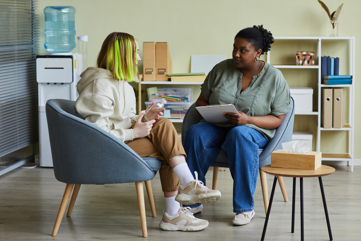 Female case manager talking to a teenager with yellow streaked hair while they sit on armchairs at office.