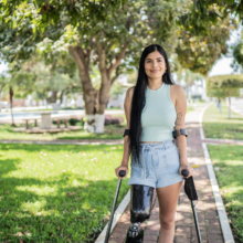 Woman with prosthetic leg stands on crutches and smiles for camera in green park.
