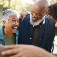 A candid outdoor photo of five adults standing close together in a circle with their arms around one another, smiling and laughing. The group includes an older woman with short gray hair wearing a denim jacket and an older man with a white beard wearing a navy blue vest. Sunlight filters through green trees in the background, giving the photo a warm, cheerful atmosphere. The focus is on joyful connection and community.