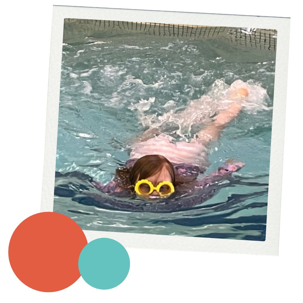 A young girl wearing a purple bathing suit and yellow googles swims in a pool during swim lessons