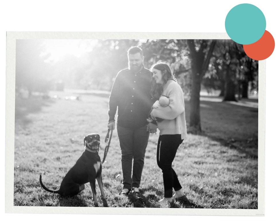 A black-and-white photo of a family walking through a sunlit park. A person walks on the left holding a dog’s leash while another person carries a small child on the right. The two adults hold hands and smile at each other. The dog sits and looks toward the camera. Decorative teal and orange overlapping dots are positioned in the upper right corner.