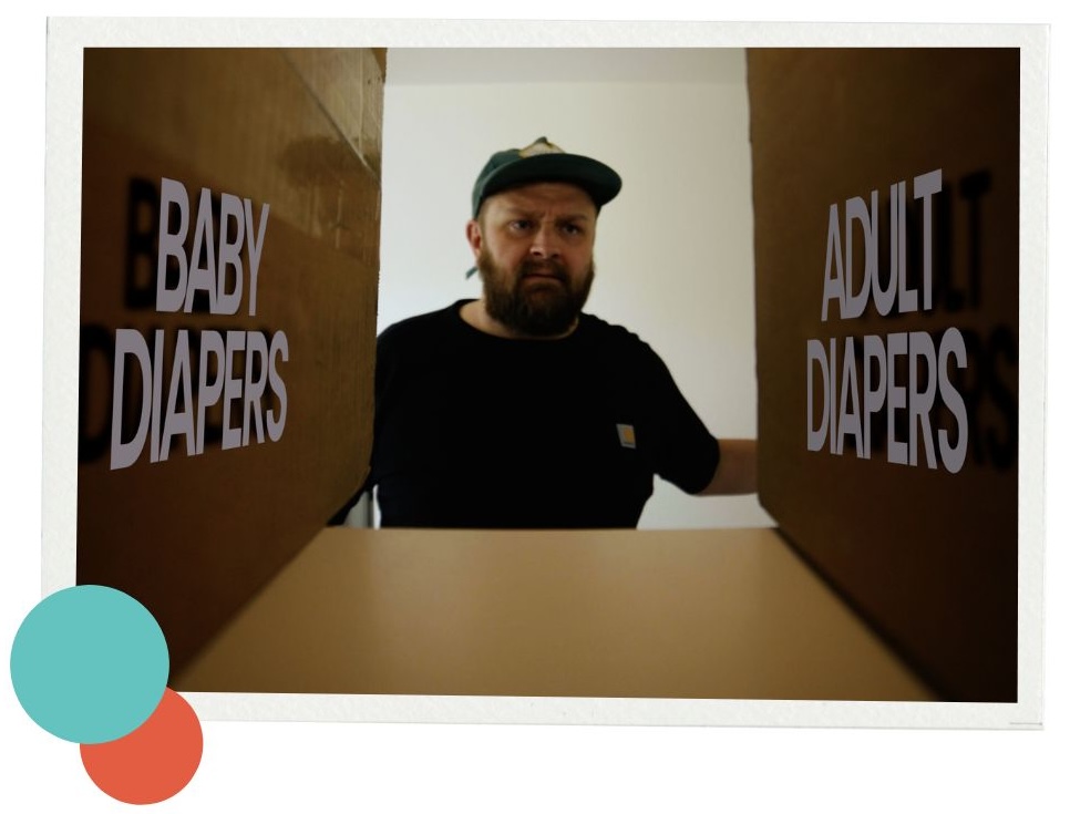 A bearded person wearing a black shirt and green cap stands between two oversized cardboard boxes, one labeled “BABY DIAPERS” on the left and the other labeled “ADULT DIAPERS” on the right. The individual appears contemplative, looking at the gap between the two boxes. In the bottom left corner, overlapping orange and teal dots add a pop of color to the white background.