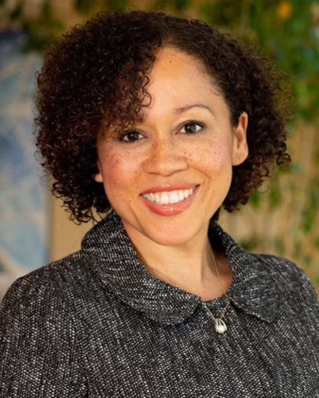 A smiling individual poses for a headshot in front of a yellow and blue background with a plant growing. The person is wearing a black suit jacket and a gold necklace.