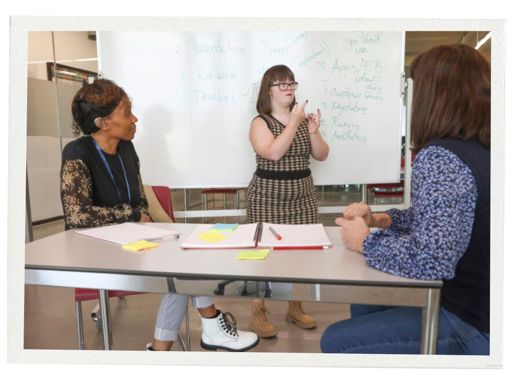 A young woman with Down syndrome gives a presentation in front of a whiteboard covered with handwritten notes. She uses sign language while two professionals, seated at a table with notebooks and sticky notes, attentively listen to her.
