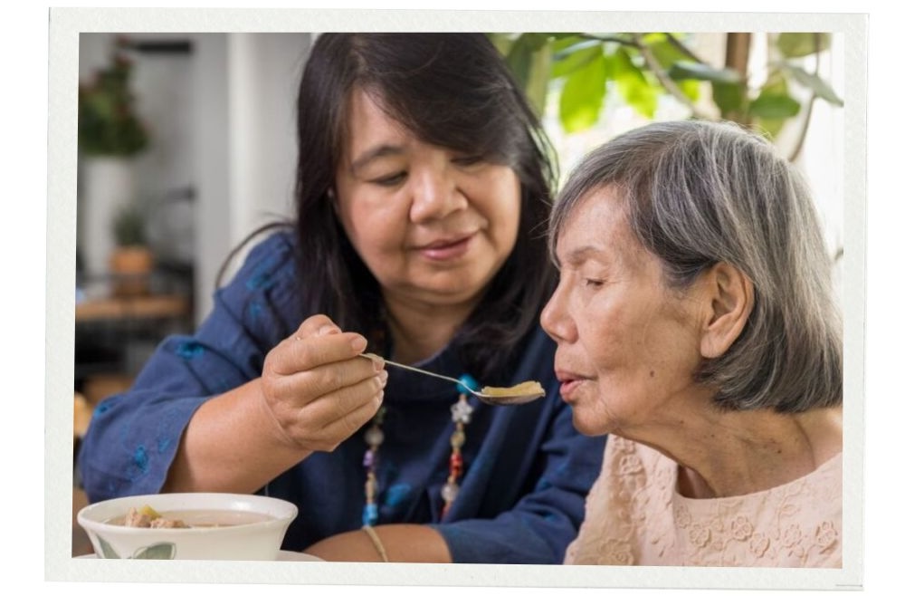 A middle-aged woman gently feeds soup to an older woman with gray hair seated at a table. The scene is warm and caring, with green plants in the background and a soft white border around the image.