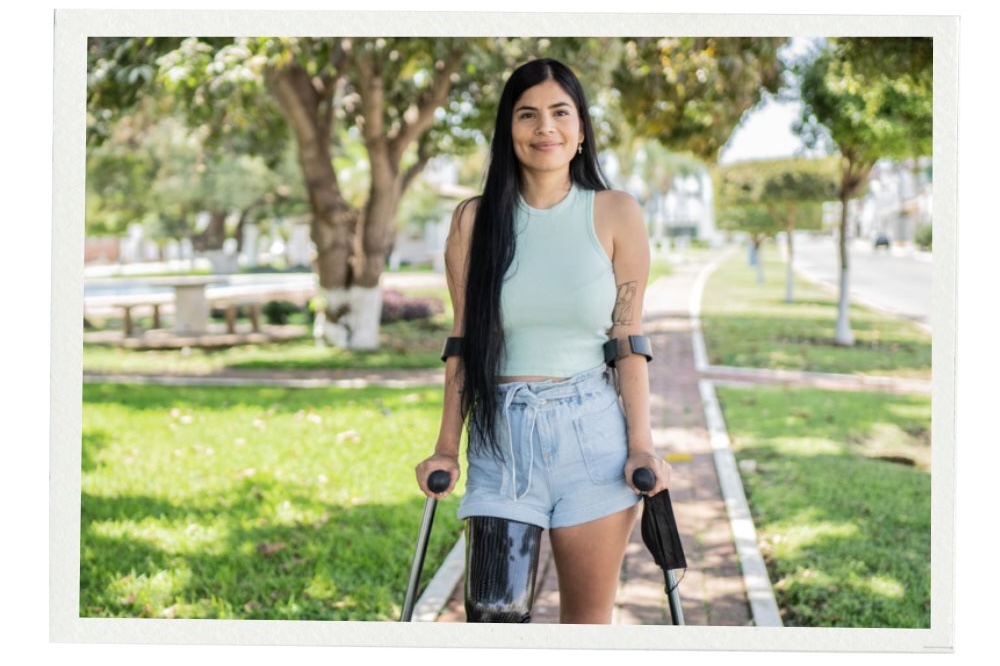 A woman with long dark hair walks confidently with forearm crutches along a sunny park path. She wears a light blue tank top and denim shorts, and her black prosthetic leg is visible. The image has a white border.