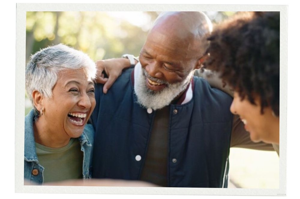 A smiling older couple and a younger adult stand close together outdoors, laughing and enjoying each other’s company. The photo has a soft white border. 