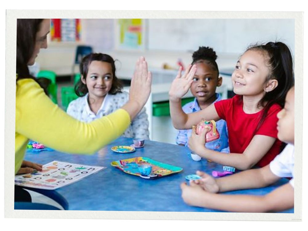 A group of four children sits at a blue classroom table. A teacher in a yellow sweater reaches out for a high five with a smiling girl in a red shirt, who is holding a toy unicorn. Other children smile and watch. The table holds colorful toys and a worksheet with large printed letters.