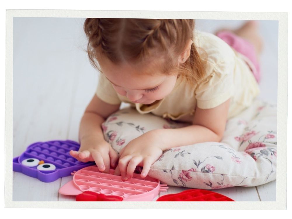 A young child with light brown hair lies on a floral-patterned cushion on a white wooden floor, focused on pressing the bubbles of a pink pop-it sensory toy. A purple owl-shaped pop-it sits nearby. The child wears a pale yellow shirt and pink shorts.