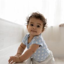 A young toddler wearing a light blue shirt with white designs stands facing the camera with a small smile.