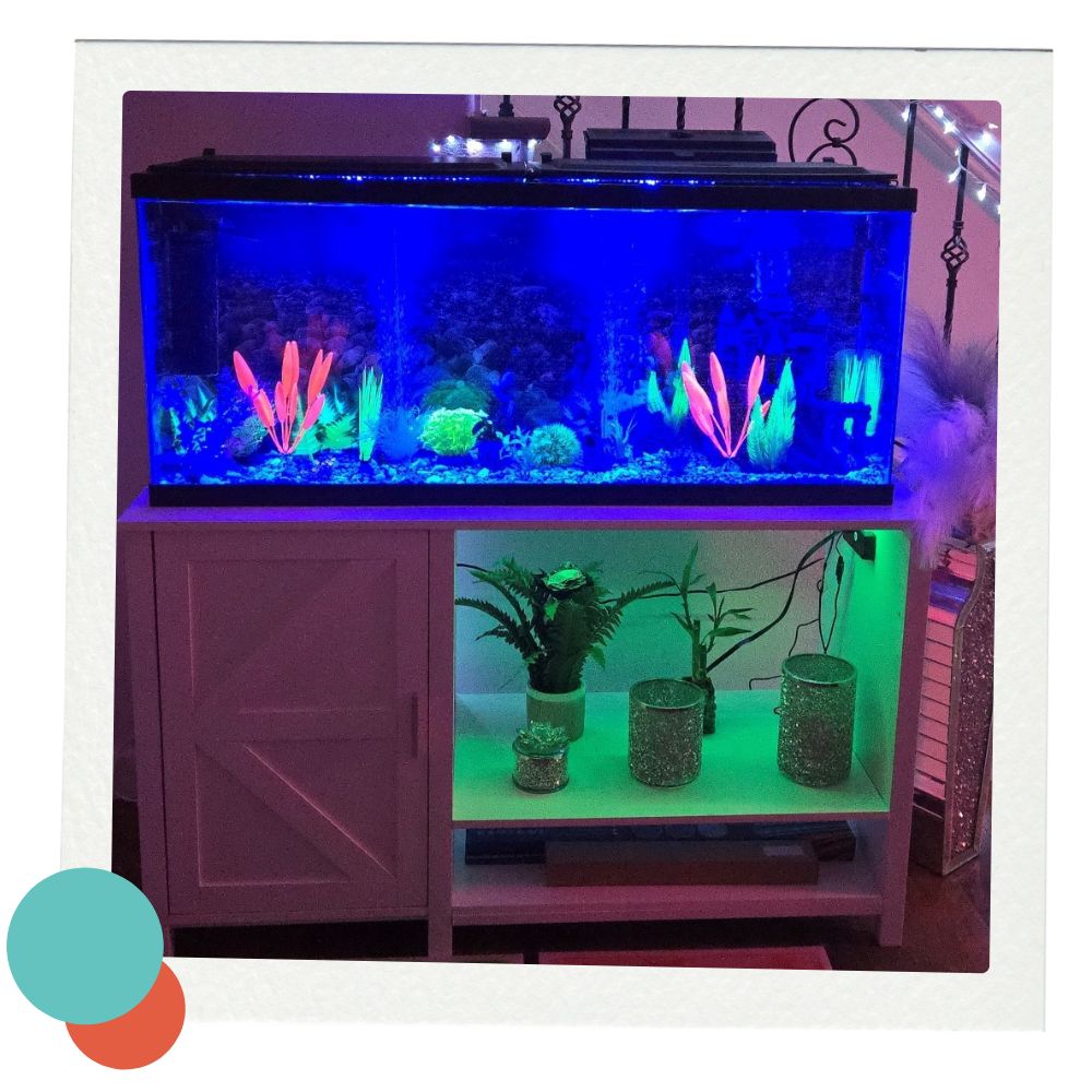 A close-up of a glowing aquarium filled with brightly colored artificial plants and pebbles. The tank sits on a white stand with open shelving. The lower shelf is lit green and decorated with faux plants and glittery silver containers. A few cables are visible in the background. Sensory tiles in shades of red and green lie on the floor in front.
