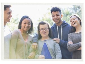 Five smiling teenagers stand together outdoors in a sunlit setting. A young woman with Down syndrome stands in the center, surrounded by friends with their arms around one another, conveying friendship, inclusion, and community. Trees and soft natural light create a bright, welcoming background.