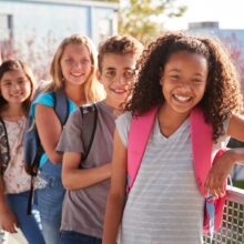 A group of four smiling school-age children wearing backpacks stands in a line outdoors, leaning against a railing. The child in front wears a gray striped shirt and a pink backpack and is smiling brightly at the camera. The rest of the group stands slightly behind in a diagonal line, with trees and a building in the background on a sunny day.