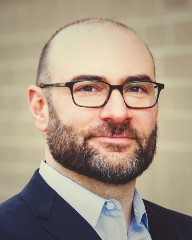 A man wearing a navy suit coat and light blue button down shirt smiles facing the camera. He has a beard and wears glasses