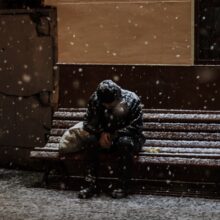 The photo shows a person sitting hunched forward on a wooden bench during snowfall at night. Snow covers their dark clothing, hat, and the bench, with flakes continuing to fall heavily. A bag sits beside them, partly dusted with snow. The scene conveys cold, isolation, and hardship, highlighting the vulnerability of people without shelter in harsh winter weather.