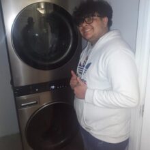 A smiling young man poses in front of a stacked washer dryer set and gives a thumbs up sign