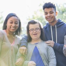 A group of five young people standing close together outdoors, smiling and laughing in bright sunlight. One person in the center wears glasses and a gray cardigan, and the others stand beside them with friendly, supportive expressions.