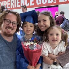 A family of four takes a selfie together at a graduation ceremony. The younger brother wears a blue graduation cap and gown and holds flowers