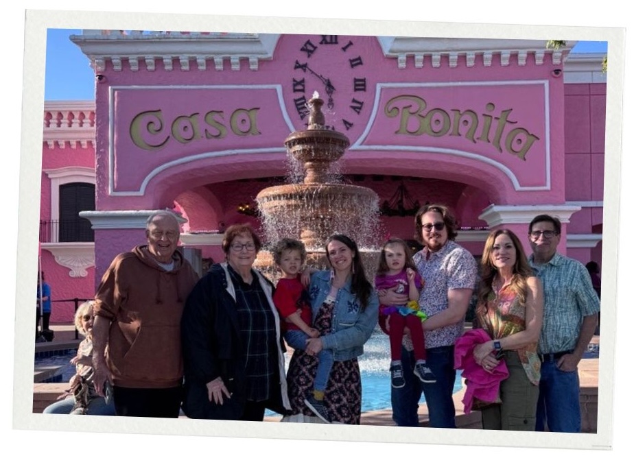 A big family of eight people poses outside of Casa Bonita, a big pink structure. The photo appears inside a polaroid frame