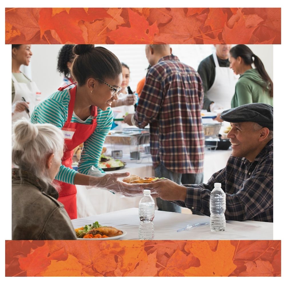 A volunteer wearing a red apron and gloves serves a plate of food to a smiling person seated at a table in a community meal setting. Other volunteers and diners are visible in the background. The image is framed with an orange autumn leaf border.