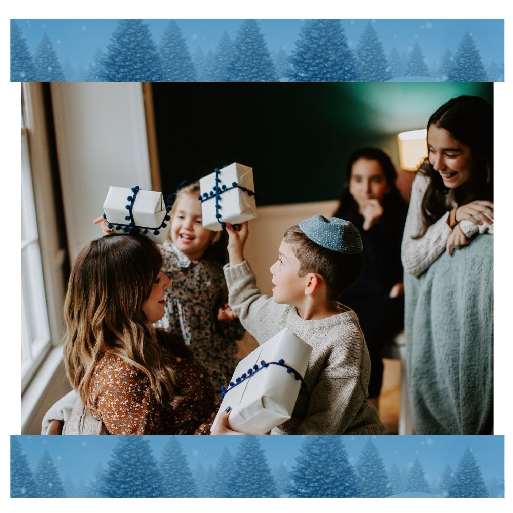 A festive photo framed with a border of blue pine trees. A group of children and adults sit together indoors near a window, smiling and playing with wrapped gifts. One child wearing a blue yarmulke and a girl in a floral dress hold small white presents tied with blue ribbon above their heads while others look on and laugh.