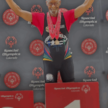 A person taking part in Special Olympics Colorado stands on the first-place podium with both arms raised in celebration. They wear several medals around their neck and a powerlifting uniform. A Special Olympics Colorado step-and-repeat banner fills the background.