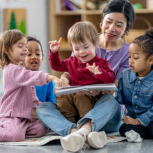 A Preschool teacher sit on the floor of the class room with children on her lap and seated beside her, as they share a story together. She is reading the children a story and they appear engaged as they listen attentively.