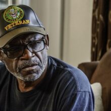 A person who is a veteran sits indoors on a couch, wearing a black U.S. Army veteran baseball cap, glasses, and a dark shirt. They look slightly to the side with a thoughtful, serious expression. Soft light comes through a nearby window and patterned curtains are visible in the background.