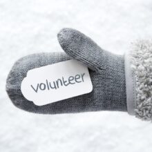 A close-up of a gray knit mitten with a white fluffy cuff, holding a white tag with the word 