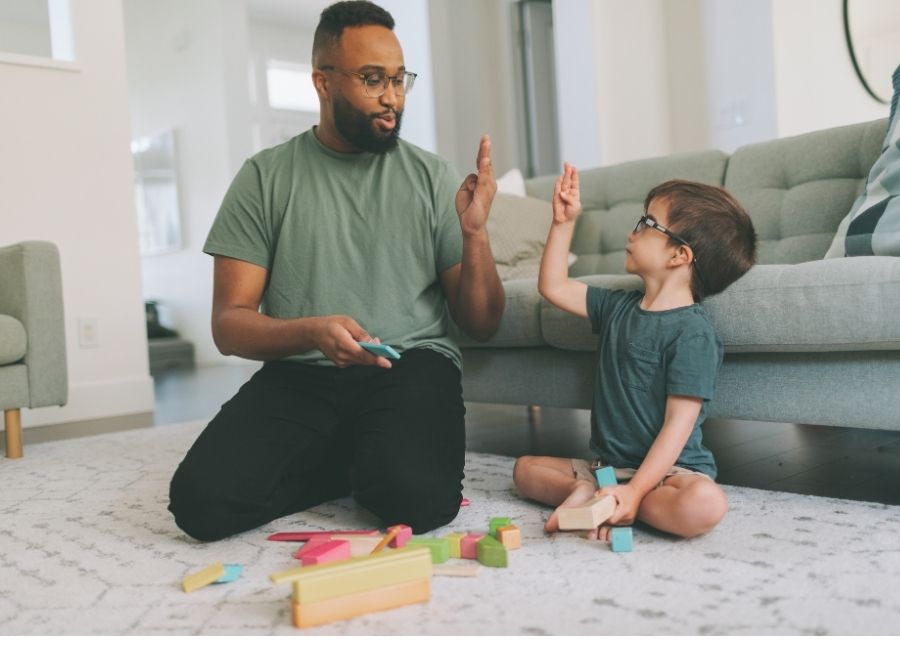 A man and young child sit on the floor of a living room practicing sign language together