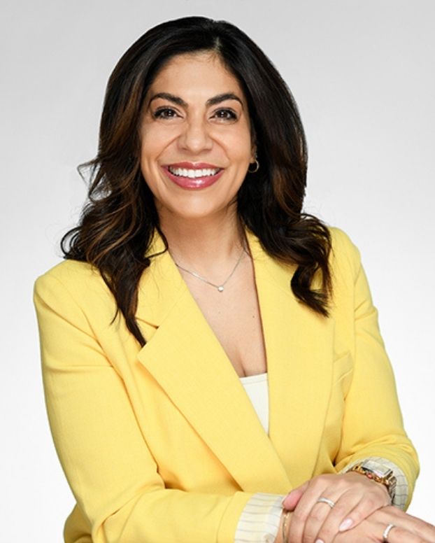 A woman with dark hair wearing a bright yellow blazer smiles at the camera in a headshot. She wears a necklace, rings and watch and has light pink painted fingernails.