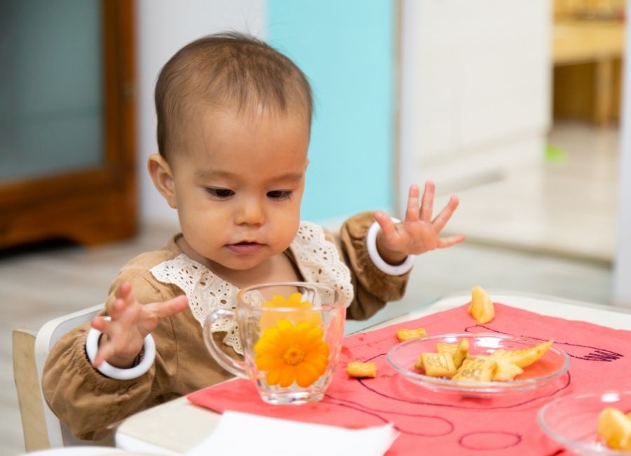 A baby sits at a table with a cup in front of them and a plate with orange food