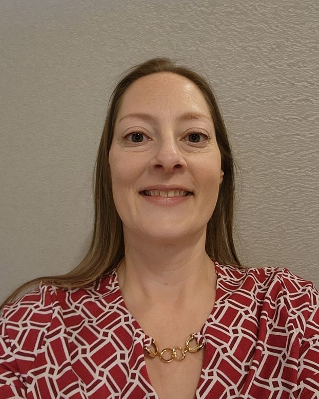 A woman smiles in a selfie against a beige wall, She is wearing a patterned red top with a gold necklace detail. She has long brown hair.