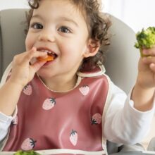 A smiling toddler sits in a high chair wearing a bib. The child has food in both hands and is eating.