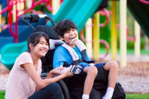 Child using a wheelchair smiles and laughs at a colorful playground alongside another child sitting nearby. Bright playground equipment in green, yellow, and pink appears in the background. The image represents inclusive summer activities and community connection, highlighting Colorado summer resources, accessible camps, recreation programs, and family-friendly opportunities for children and youth across the Denver metro and statewide in Colorado.