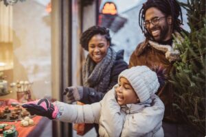Family bundled in winter coats, scarves, and hats smiles while looking at a holiday storefront display. A child in a knit hat and gloves reaches toward festive decorations in a shop window, while two adults stand nearby holding a small evergreen tree. The image represents statewide holiday support resources in Colorado, including holiday assistance programs, toy drives, meal programs, winter gift support, and community events available to individuals and families across urban and rural communities.