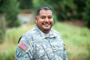 Portrait of a smiling U.S. military service member in uniform standing outdoors with greenery in the background. An American flag patch is visible on the sleeve. The image represents support for veterans and military families across Colorado’s Front Range, including resources in the Denver metro, Colorado Springs, and surrounding communities. It reflects access to housing assistance, employment services, health care, and community programs tailored to veterans and active-duty military members throughout the region.