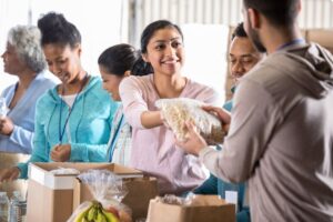 Volunteers distribute food at a Colorado food bank, handing a bag of dry goods across a table filled with boxes and fresh produce. A diverse group of adults stands indoors in a community setting, smiling and organizing groceries for families in need. The image represents food access support in Colorado, including food banks, food distribution programs, and community hunger relief services in the Denver metro and Colorado Springs areas.