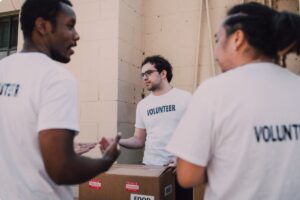Three volunteers wearing white shirts labeled “VOLUNTEER” stand outdoors beside a building, talking while organizing a large cardboard box marked for food distribution. The image represents volunteer opportunities across Colorado’s Front Range, highlighting ways community members can give back.