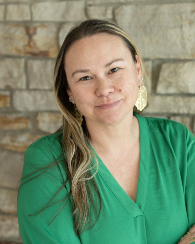 1 Ki'i Powell, RMHS Chief Operations Officer. A woman with long brown and blonde highlighted hair, wearing a green v-neck top and gold leaf earrings, smiling with arms crossed in front of a stone wall.