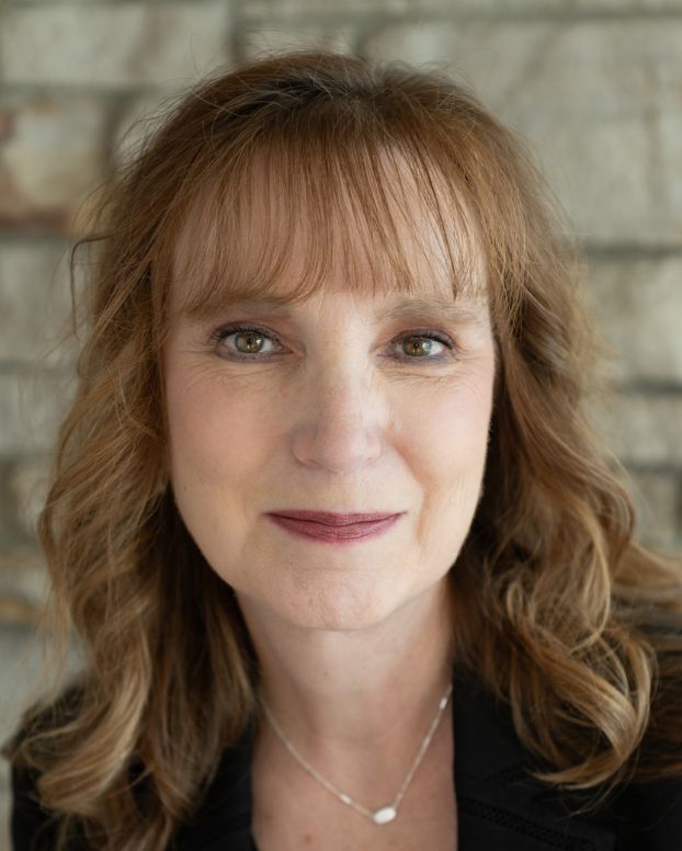 3 Nancy Stokes, RMHS Chief Financial Officer. A woman with long, wavy auburn hair and bangs, wearing a black top and a delicate silver necklace, smiling in front of a stone wall.
