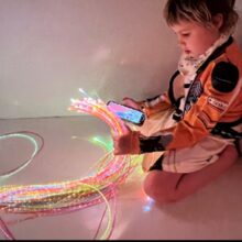 A child sits on the floor holding a bundle of fiberoptic lights in one hand and a cell phone in the other