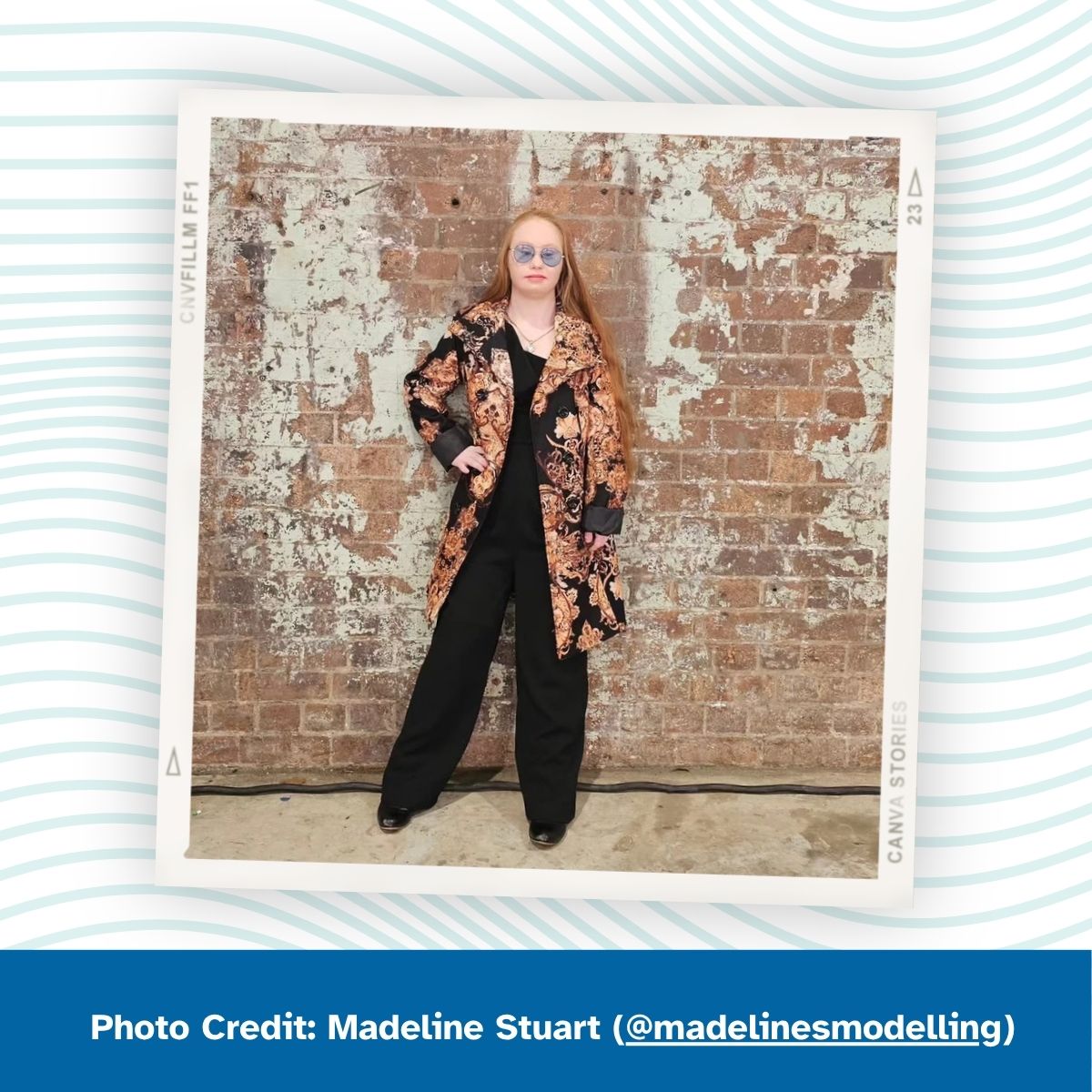  A woman with Down syndrome stands in front of a weathered brick wall. She wears a black outfit with a black and gold patterned coat, wide-leg pants, and round blue sunglasses, with long red hair. Her hand rests on her hip. Photo Credit: Madeline Stuart (@madelinesmodelling)