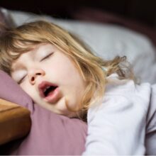 A young child with blonde hair sleeps on their side, resting their head on a pillow next to a wooden bedside table, mouth slightly open in a peaceful expression.