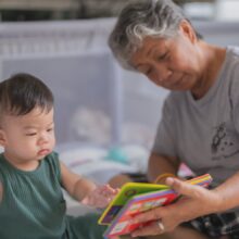 An older adult sits with a young child, reading a colorful board book together in a cozy indoor setting. The child reaches toward the book while both focus on the pages, creating a calm and engaging learning moment.