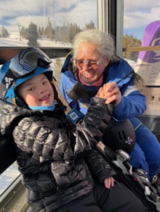 A smiling child wearing a blue ski helmet, black puffer jacket, and ski gear sits inside a ski gondola, holding hands with an older adult in a blue ski jacket. Both are smiling warmly at each other, with snow-covered trees and mountains visible through the gondola windows in the background.