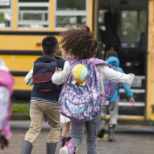 Children wearing colorful backpacks walk toward and board a yellow school bus. The image is photographed from behind, capturing students heading to school, with the bus doors open and other children visible in the background.